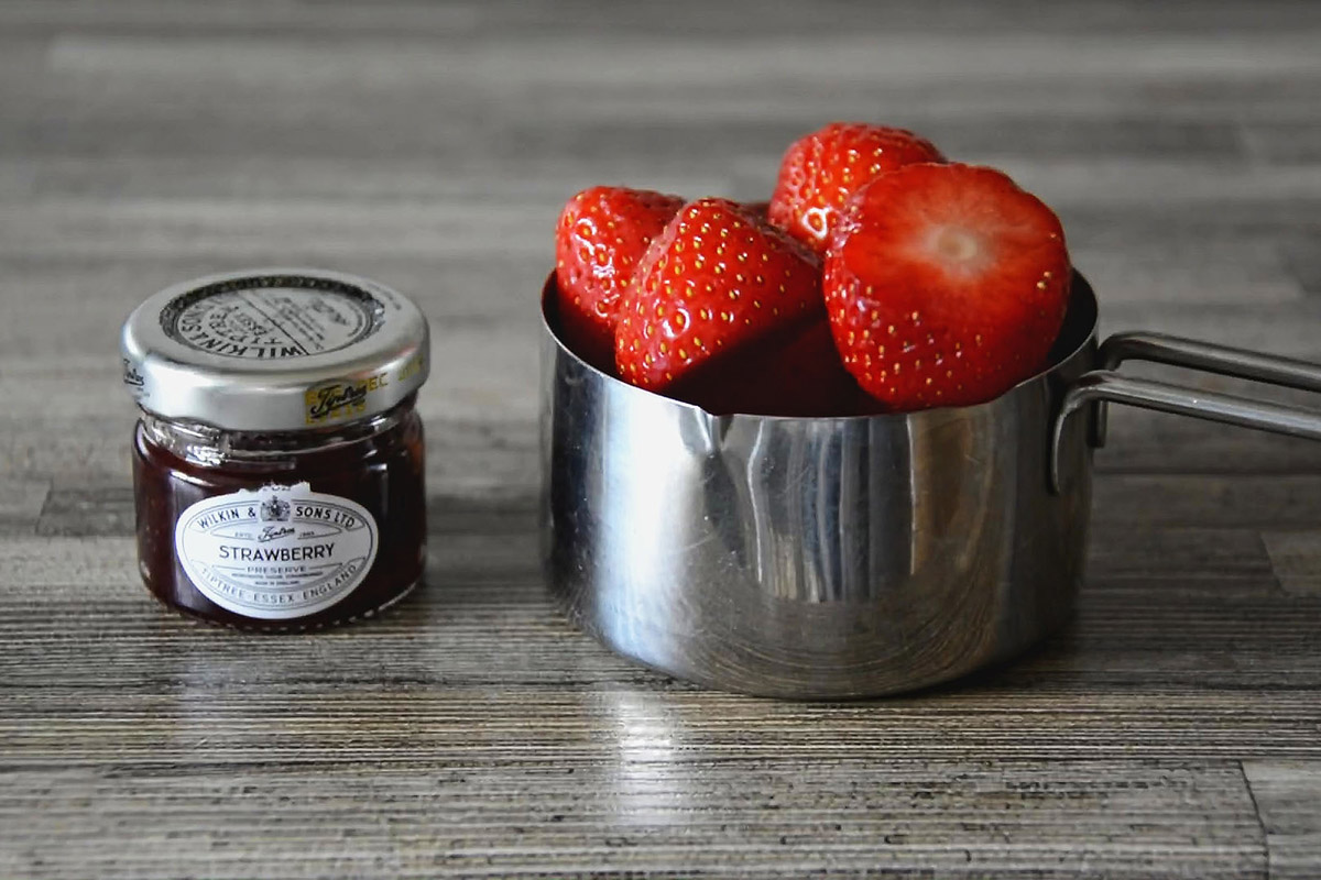 Miniature jar strawberry jam besides steel measuring cup full of fresh strawberries