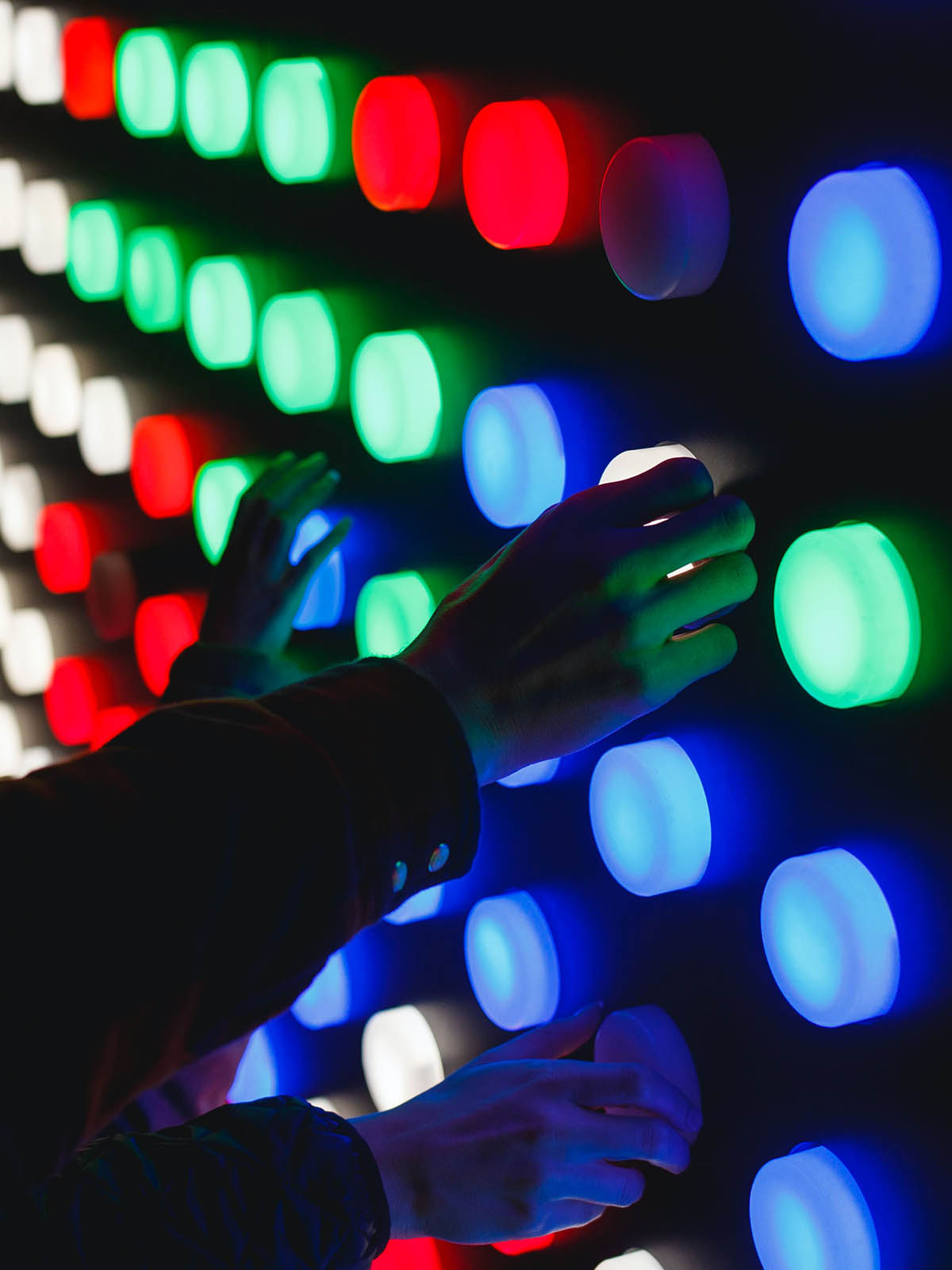 Three right hands from three different people pressing large multicoloured, illuminated buttons set in rows on a black wall.