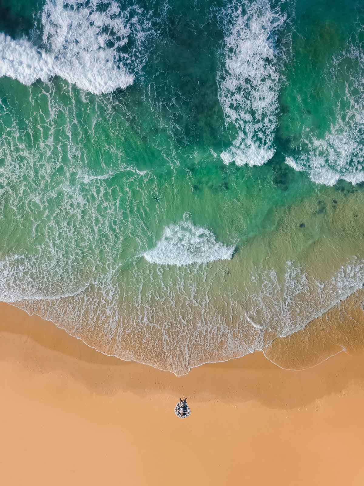 Bird’s eye view of a person lying at the edge of a golden sandy beach with turquoise waves breaking on the shoreline.