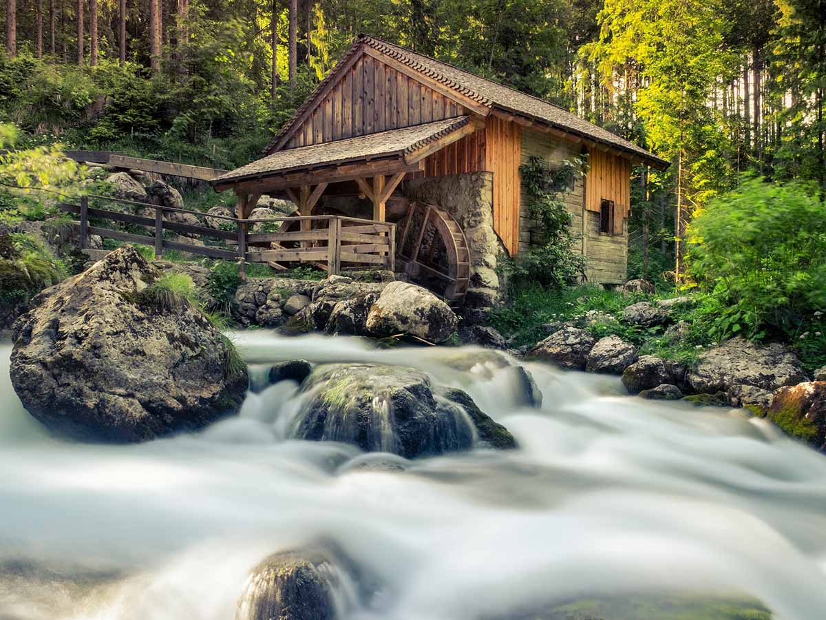 Idyllic stone and wood cabin with water wheel behind blurred water, flowing between mossy rocks.