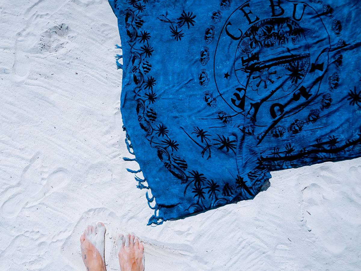 Looking down at feet in powder white sand. Tasseled blue sarong with black palm tree print laid out on sand.