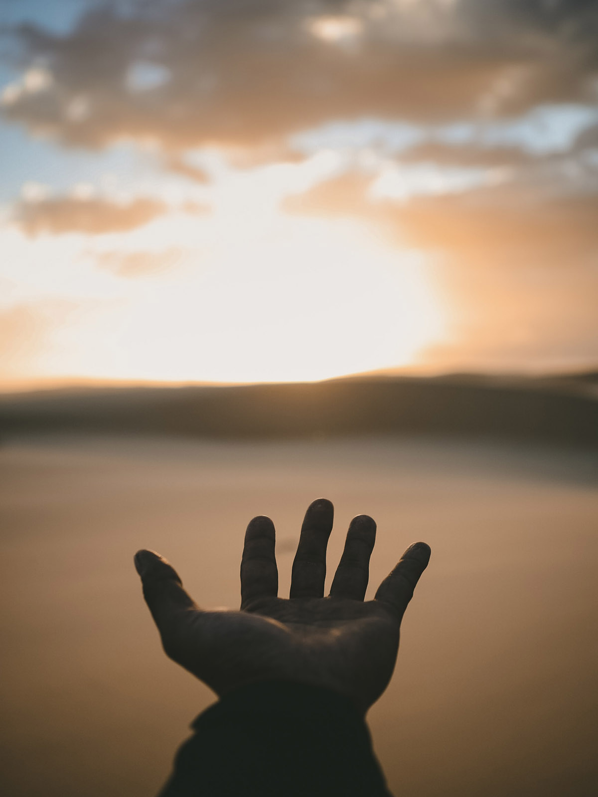 Silhouetted outstretched hand reaching across the beach towards a distant sunset
