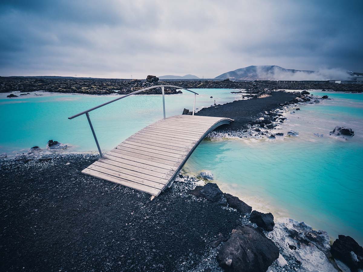 Weathered wood bridge connecting small black lava sand islands amongst ice blue glacial hot pools.