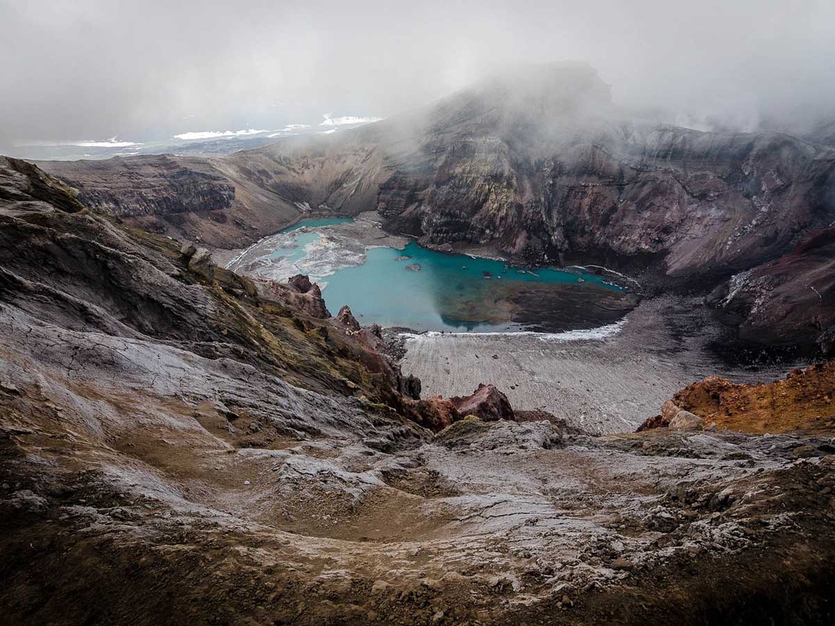 View into volcanic crater towards glacier blue lake below. Rising steam and smoke from hot water.