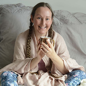 Michelle, the irritable vegan, wears a pale pink, cosy shawl and pigtails whilst holding a copper mug of frothy coffee between both hands and smiling at the camera.