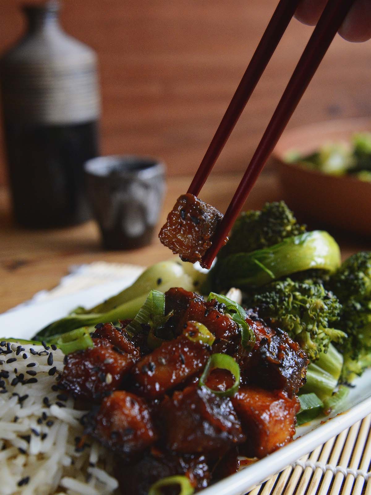 A cube of sticky, marinated tofu being lifted from the plate by a pair of red chopsticks. The plate contains a mound of glazed tofu besides white rice and green vegetables, garnished with black sesame seeds.