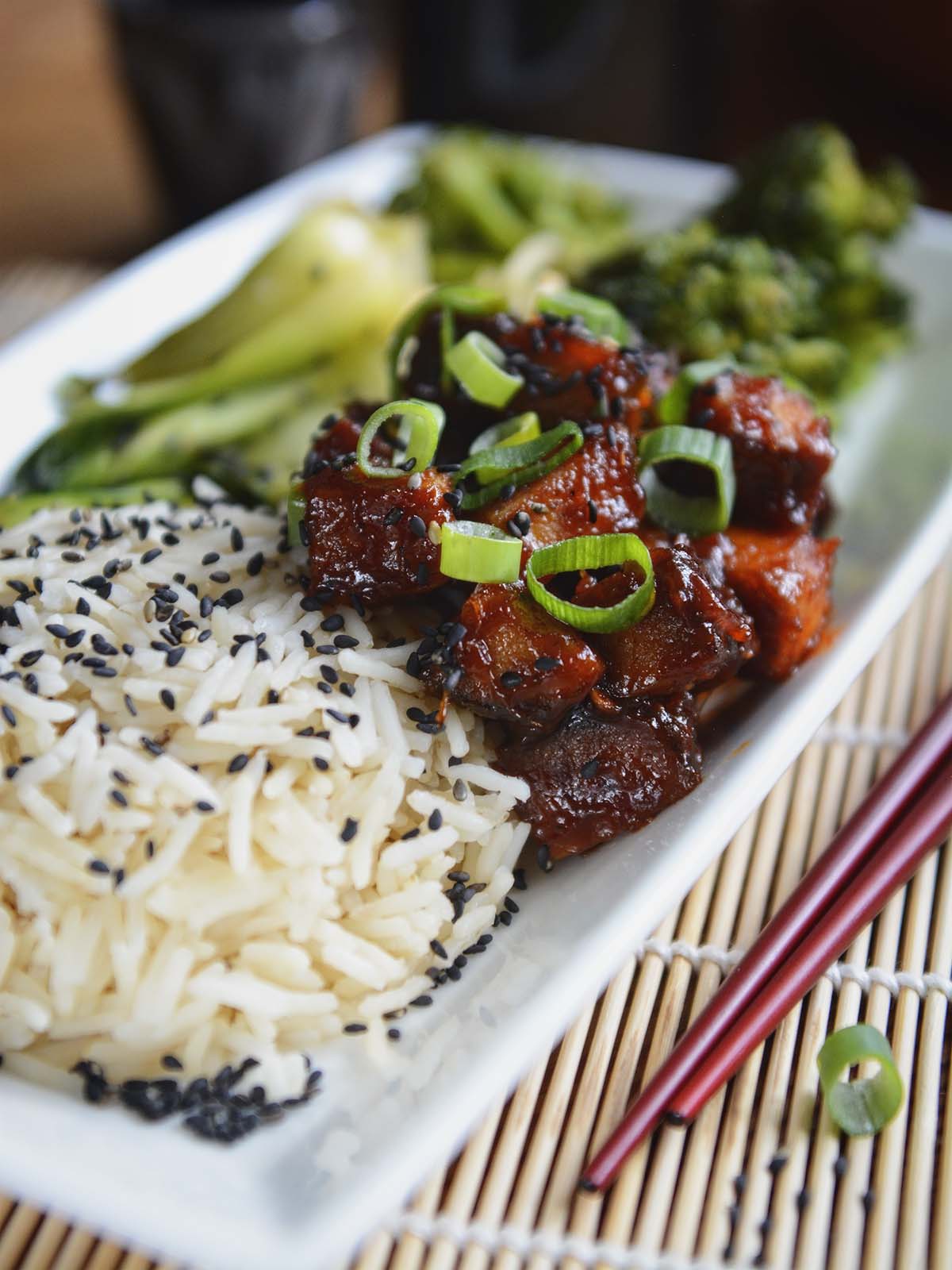 Cubes of sticky tofu, white rice and green vegetables, garnished with green onions and black sesame seeds, presented on a white rectangular plate.