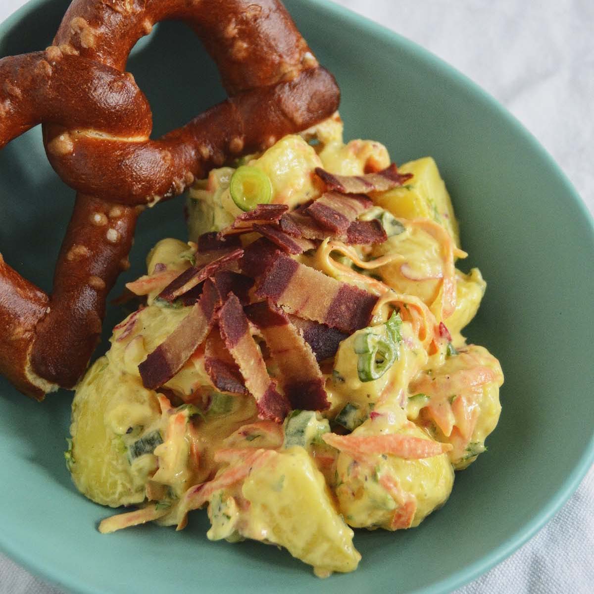 Creamy potato salad topped with chopped bacon and a baked soft pretzel.