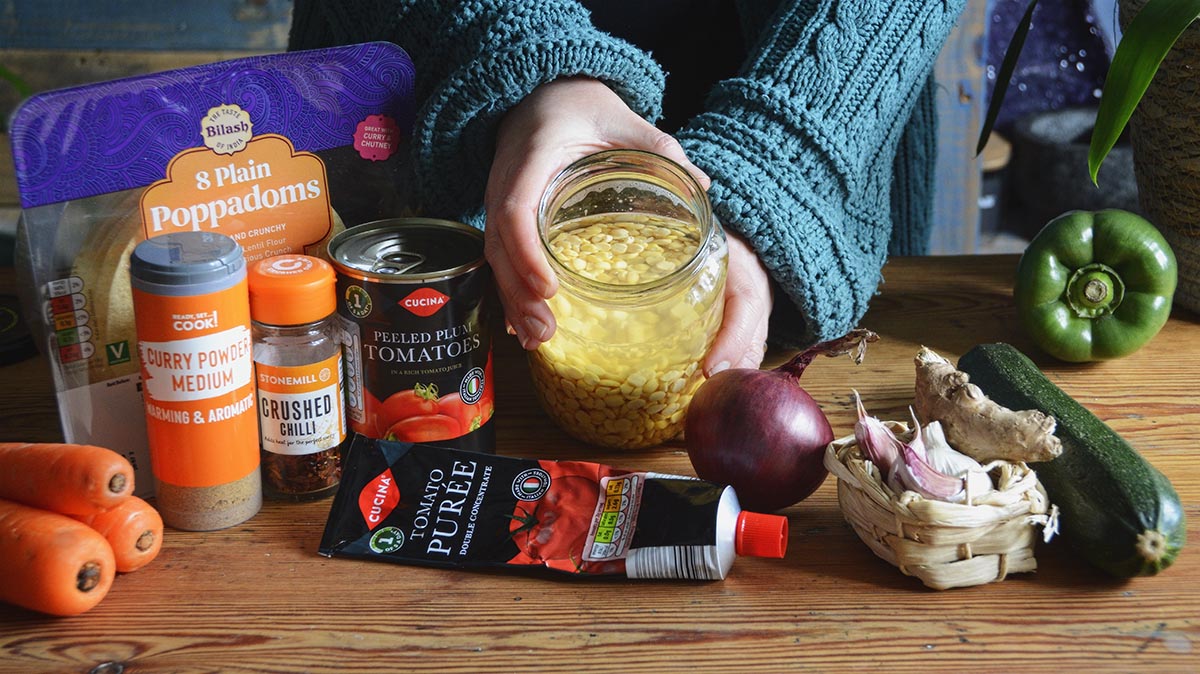 An array of ingredients to make a lentil dahl displayed on a wooden tabletop. A glass jar of soaked chana dahl is held between two hands.