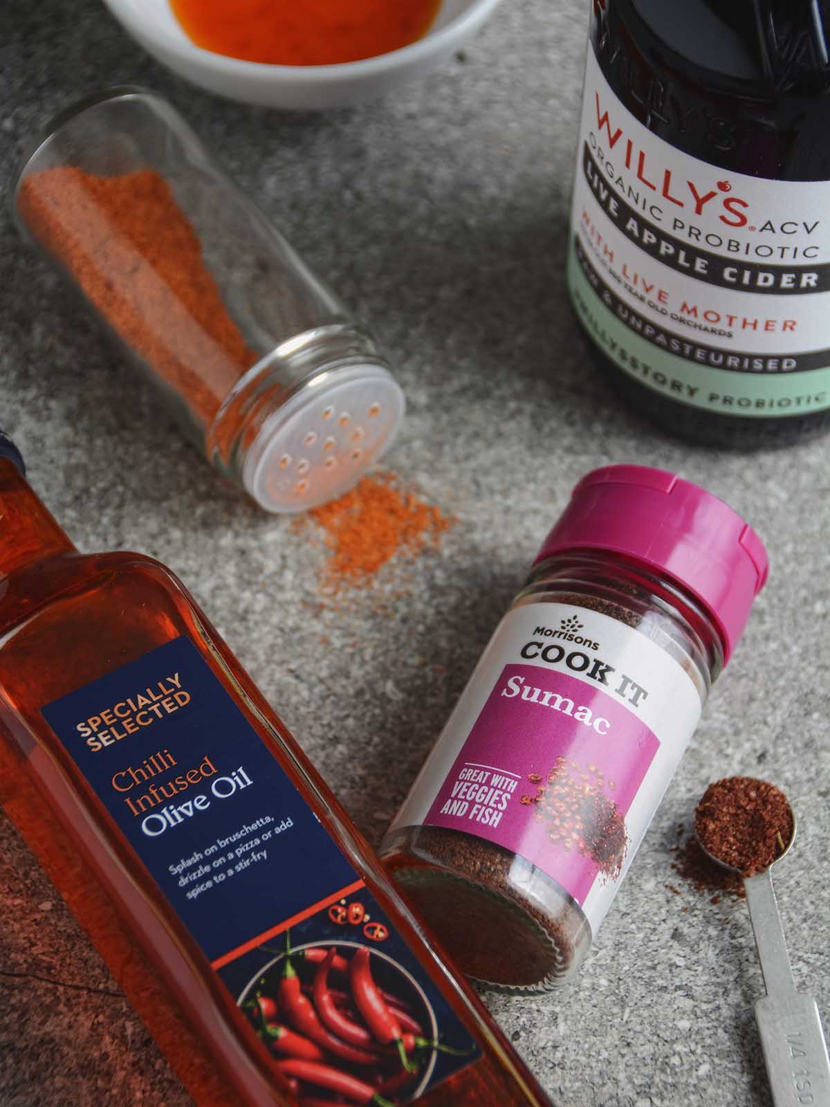 The salad dressing ingredients arranged on a grey background. The vibrant orange-red chili oil, sumac in a measuring spoon next to its spice jar, and an uncapped jar of peri peri salt with some spilled spice. Alongside them, a bottle of Willy's brand ACV stands upright, while the other ingredients are neatly laid out.