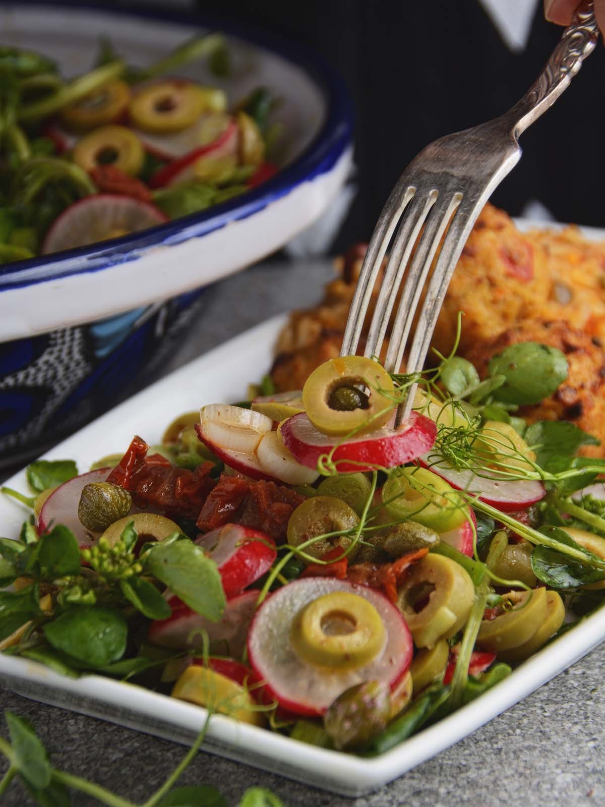 A forkful of the radish and olive salad is being lifted from the plate. The vibrant colors of the salad ingredients, including sliced red radishes, juicy green olives and wispy pea shoots are visible.