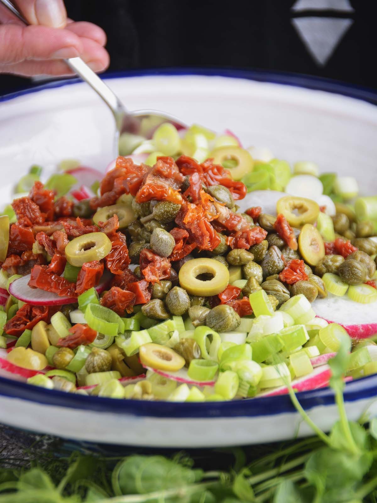 The radish and olive salad ingredients are mounded up in a white salad bowl where they have been thrown in, one atop the other. In the background a silver spoon is just beginning to lift the ingredients in order to combine them into a tossed salad.