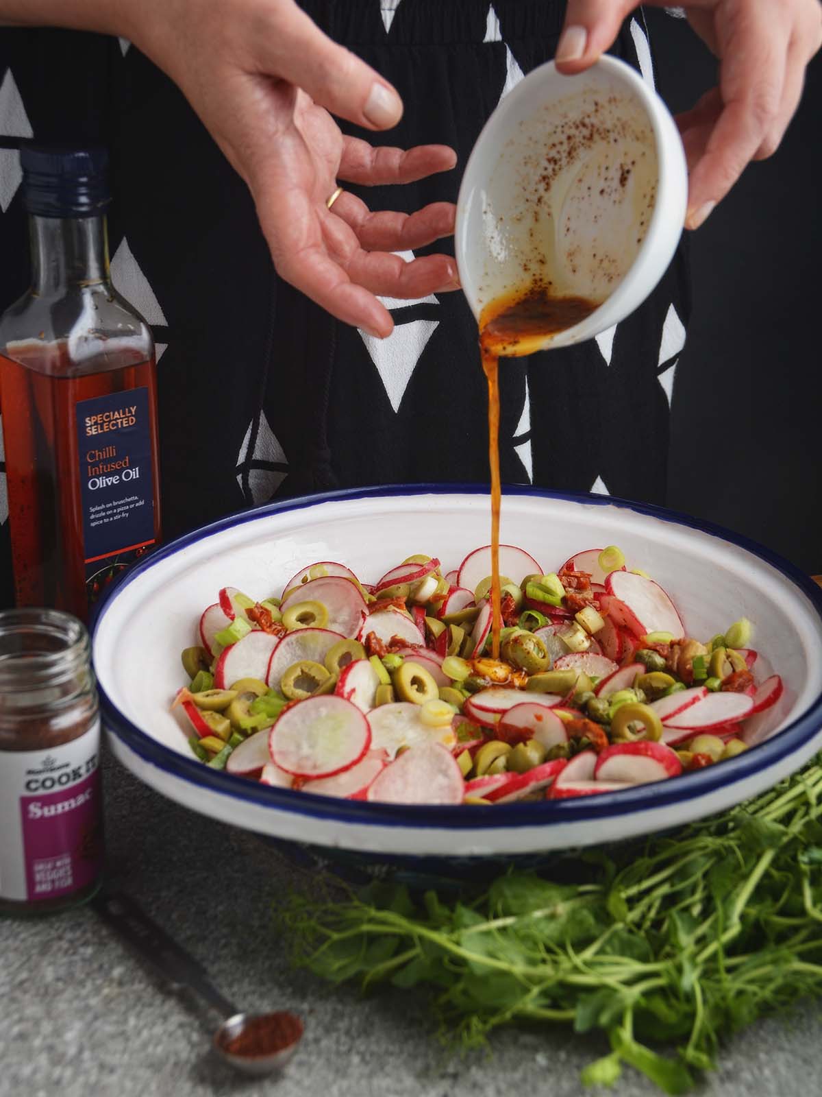 Hands pouring a steady stream of chili oil dressing onto the radish and olive salad. The dressing is being poured from a small white bowl with a visible coating of sumac spice. Beside the salad bowl, there is a glass bottle of olive oil and a jar of sumac. In front there is a measuring spoon filled with sumac and leafy green pea shoots.