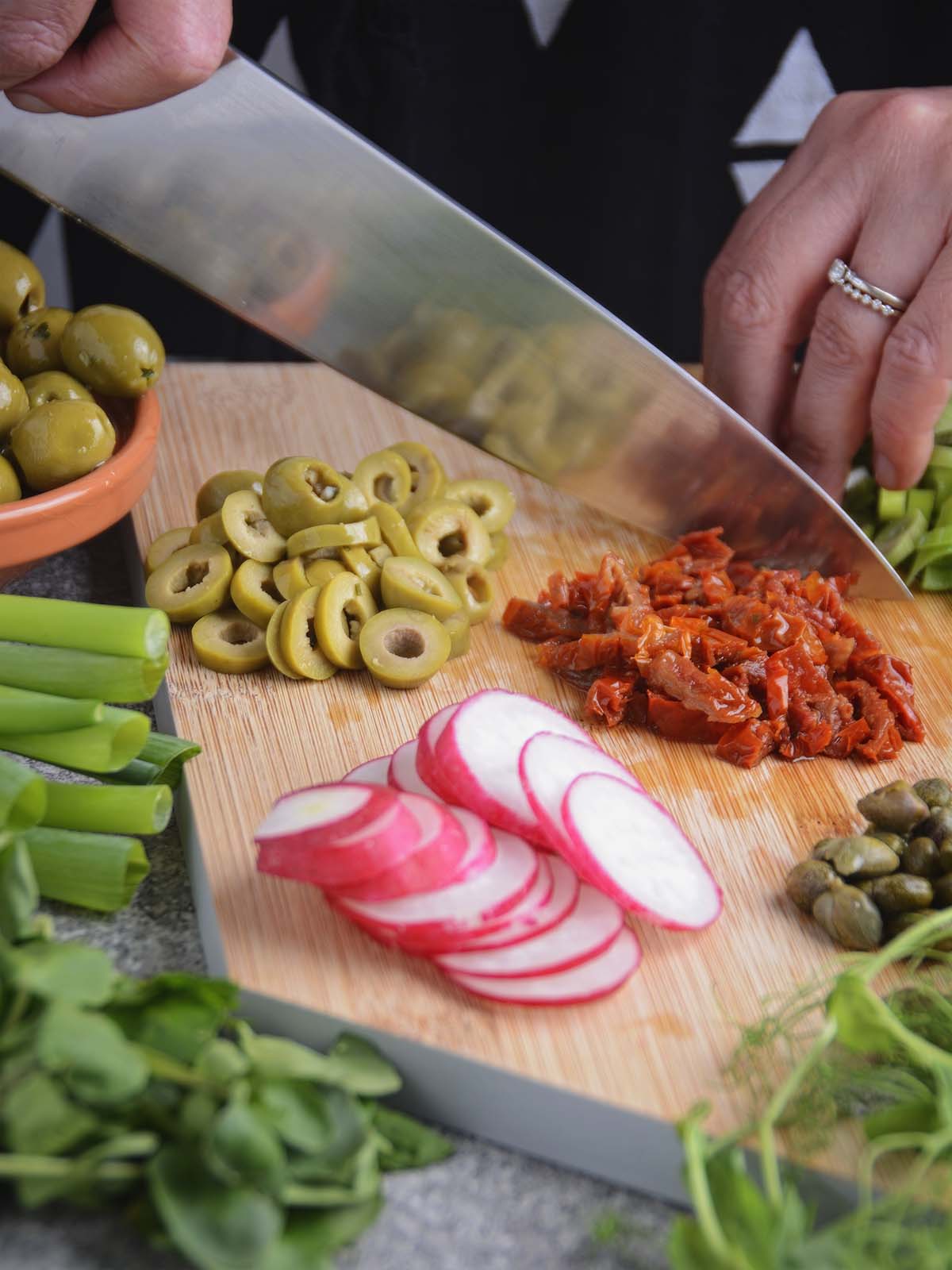 A sharp chef's knife cutting up sundried tomatoes for the salad on a wooden chopping board. The board also contains thinly sliced red radish, green olives and capers. Whole ingredients are arranged around the board, awaiting their turn to be chopped.