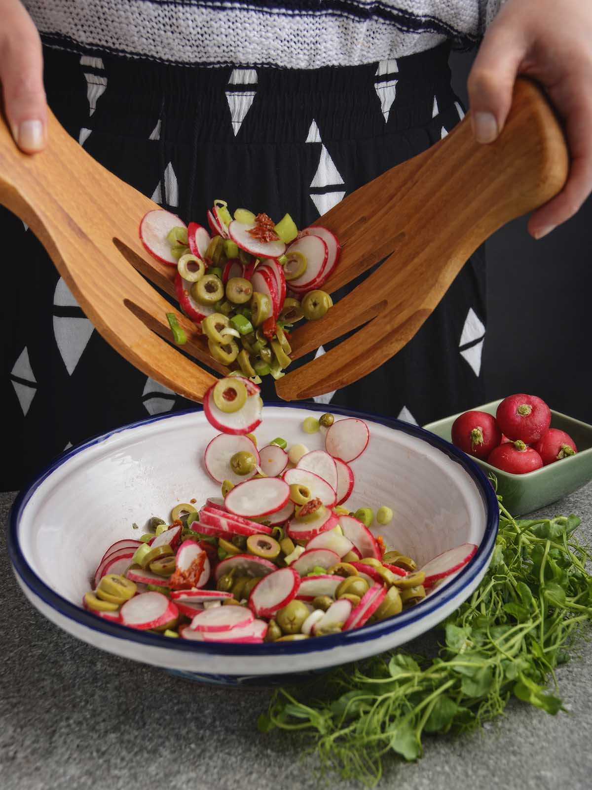 The vibrant radish and olive salad ingredients are tumbling between two wooden serving forks into a white pot bowl with a dark blue rim. The hands holding the forks are moving up out of view as the salad falls down.