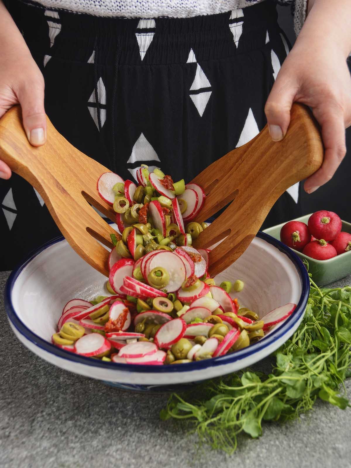 Hands tossing the radish and olive salad between two wooden salad forks. The vibrant salad ingredients are suspended between the two forks above a white pot bowl with a dark blue rim. The pea shoots lay in front of the bowl and have not yet been added.